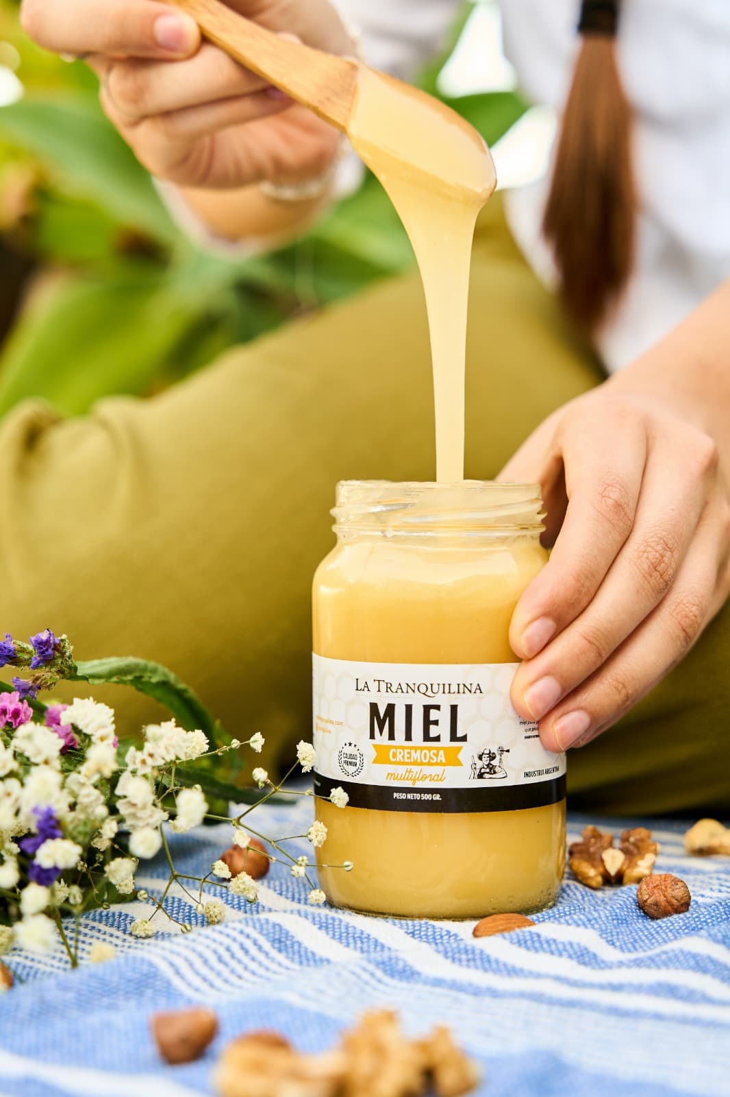 Manos sirviendo miel cremosa La Tranquilina con una cuchara de madera, rodeada de flores y frutos secos